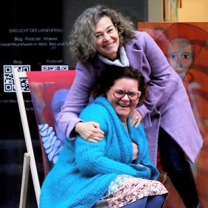 zwei lachende Frauen vor einem Schaufenster. Eine sitzt im Liegestuhl mit einem Bildmotiv bespannt, die andere beugt sich stehend dahinter nach vorne um umfasst ihre Schultern.