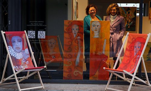 zwei Frauen mit Blick nach vorne stehen in einem Schaufenster mit 2 hochformatigen gemalten Bildern davor. Links davon ein Liegestuhl mit einem Bildmotiv bespannt. Auf dem Gehsteig davor stehen rechts und links der Frauen nochmal zwei weitere Liegestühle